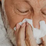 Starszy mężczyzna zamyka oczy, wycierając nos chusteczką. Widać oznaki chorób i troski.