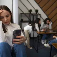 Dziewczyna z fobią społeczną siedzi sama, trzymając telefon, podczas gdy inni rozmawiają w tle.