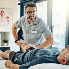 Fizjoterapeuta wykonuje świadczenie rehabilitacyjne, pomagając starszemu pacjentowi w ćwiczeniach nogi.