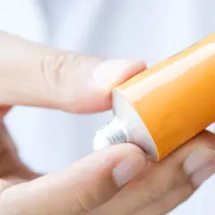 Dłonie aplikują maść z antybiotykiem z pomarańczowej tubki na palec.