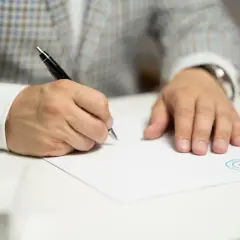 Mężczyzna w eleganckim garniturze podpisuje umowę zlecenia. Długopis w dłoni, biała kartka i pieczęć firmy.