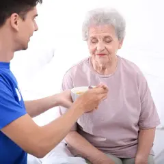Młody mężczyzna karmi starszą kobietę zupą. Dbanie o seniorów, nawet tych zmagających się z otyłością, jest ważne.