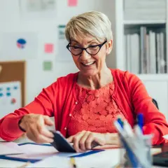Uśmiechnięta starsza pani przy biurku sprawdza coś na telefonie, zastanawiając się, ile można dorobić do emerytury.