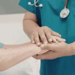 Dłoń pielęgniarki obejmuje dłoń pacjenta, symbolizując wsparcie i troskę. To obraz, który może kojarzyć się z potrzebą uzyskania **zasiłku pielęgnacyjnego**.