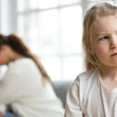 Smutna dziewczynka patrzy w bok, podczas gdy jej matka w tle ukrywa twarz w dłoniach. To obrazek pokazujący skutki parentyfikacji.