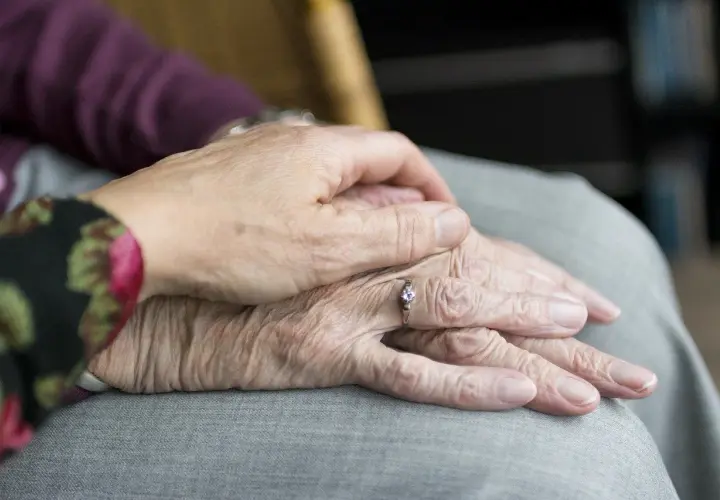 Dłonie starszej osoby, z widocznymi zmarszczkami i pierścionkiem, spoczywają na kolanach. Młodsza dłoń delikatnie je obejmuje, dając poczucie bezpieczeństwa i troski, jakby łagodziła każdy ruch, niczym lek akineton.