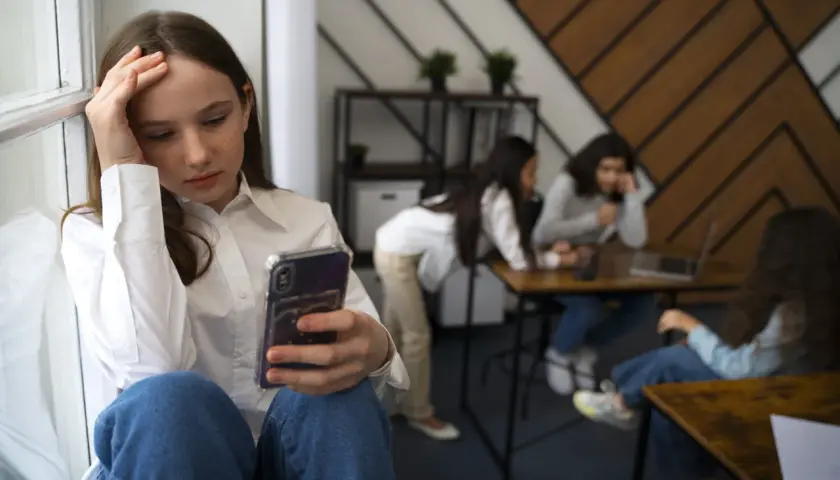 Dziewczyna z fobią społeczną siedzi sama, trzymając telefon, podczas gdy inni rozmawiają w tle.
