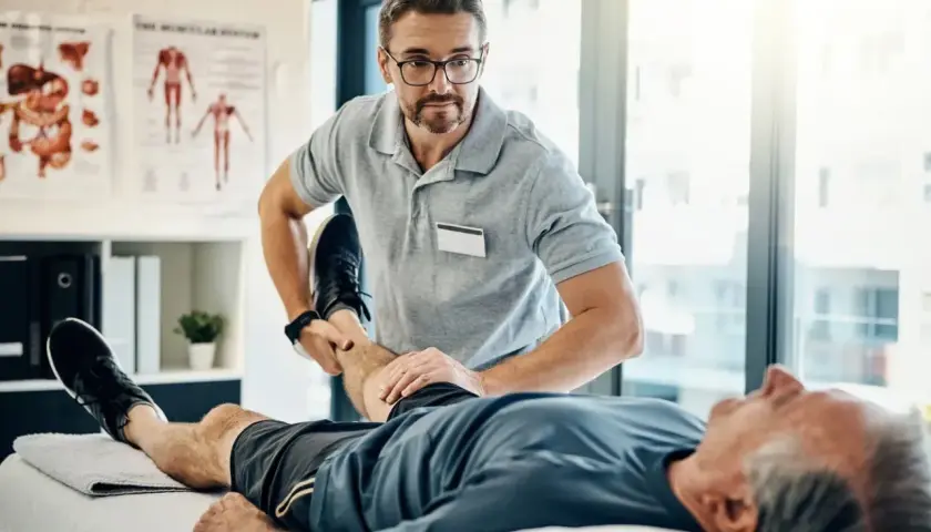 Fizjoterapeuta wykonuje świadczenie rehabilitacyjne, pomagając starszemu pacjentowi w ćwiczeniach nogi.
