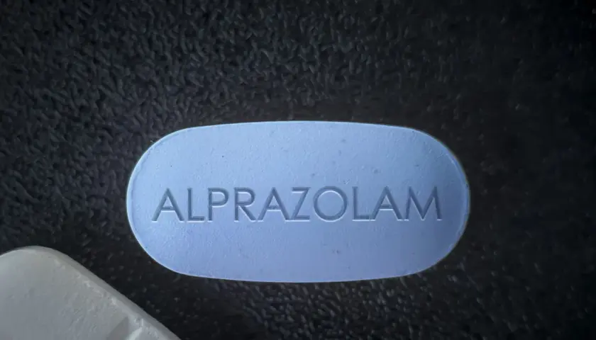 Niebieska tabletka z napisem ALPRAZOLAM leży obok białej tabletki na ciemnym, teksturowanym tle.