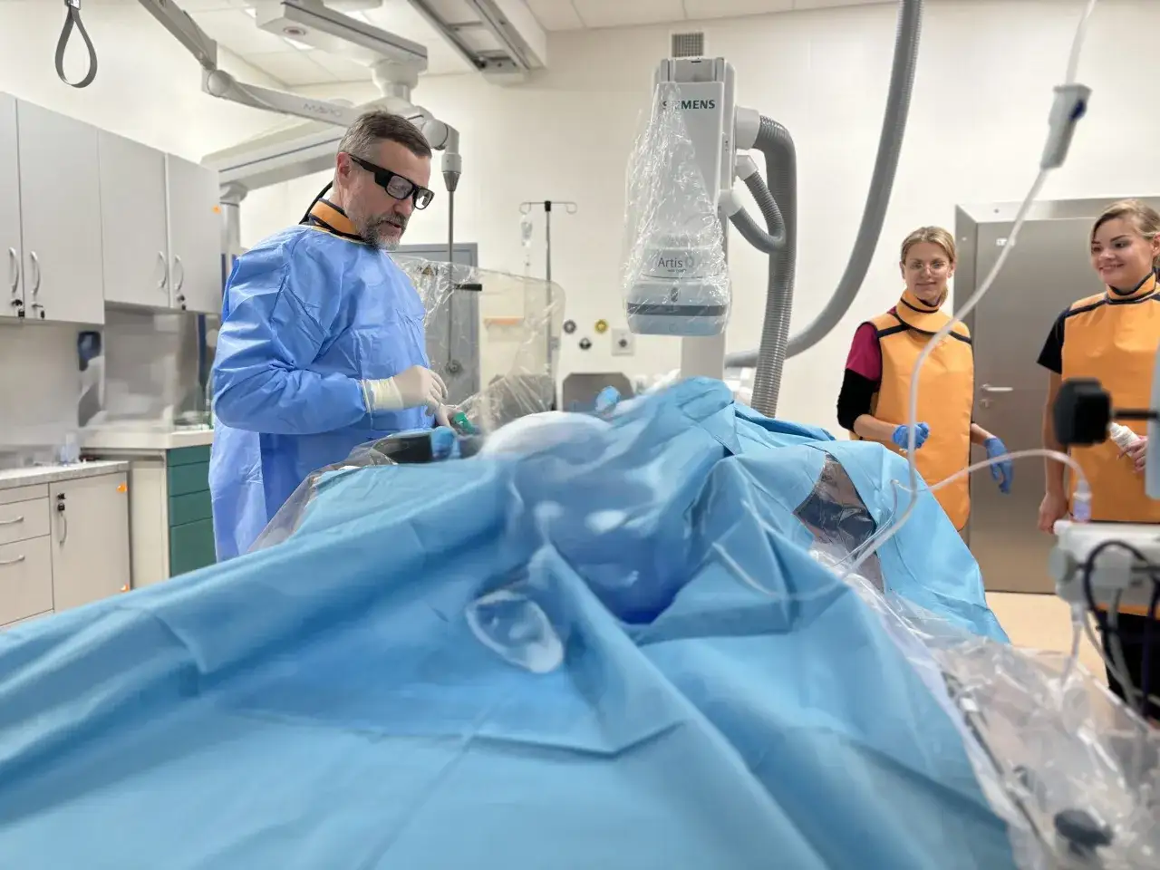 Zespół medyczny przygotowuje się do zabiegu koronarografii. Lekarz w niebieskim fartuchu i dwie pielęgniarki w fartuchach ochronnych pracują przy pacjencie.