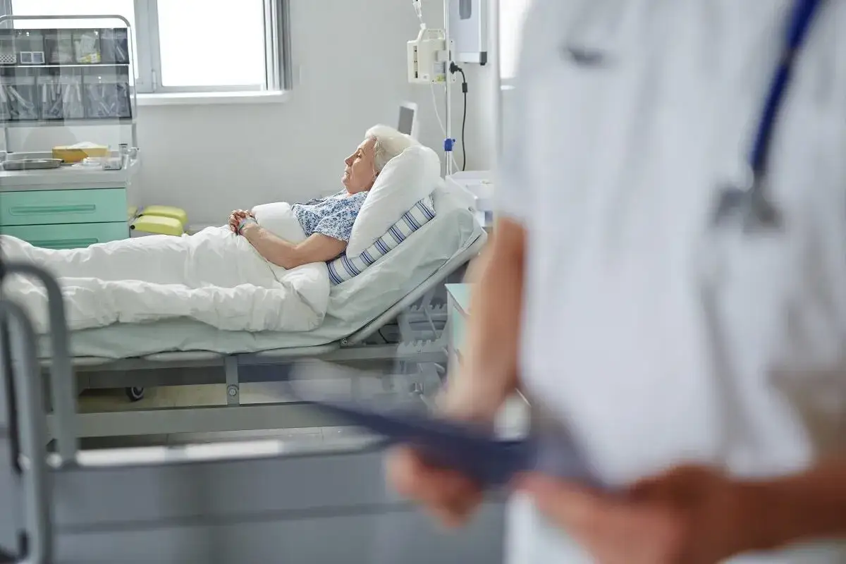 Starsza pacjentka leży w łóżku szpitalnym, otoczona opieką. To obraz troski w geriatrii.