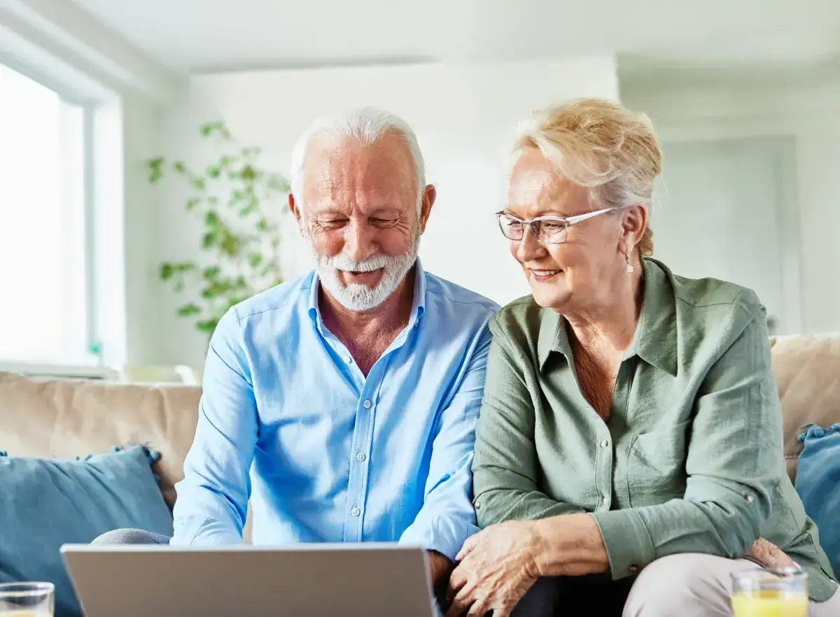 Uśmiechnięci seniorzy przeglądają coś na laptopie. Mężczyzna w niebieskiej koszuli i kobieta w zielonej bluzce siedzą na kanapie. To dla nich jak pit 0, bo świetnie sobie radzą z technologią.