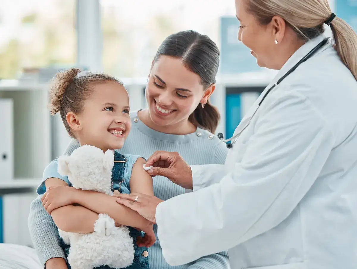 Uśmiechnięta dziewczynka z misiem dostaje szczepienie. Mama ją wspiera, a lekarka dba o opiekę nad zdrowym dzieckiem.